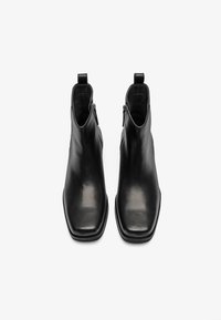 Botas de cuero negro hasta el tobillo con punta cuadrada, textura suave, cremalleras laterales y tiradores para facilitar su uso. Diseño simple y moderno.