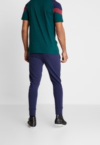 Korte mouwen shirt in groen met bordeauxrode en marineblauwe schouderpanelen, gecombineerd met marineblauwe joggers en zwarte sportschoenen. Gladde stof, nauwsluitend ontwerp.