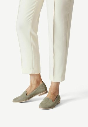 Unterschenkel in off-white maßgeschneiderten Hosen und hellgrünen Wildlederslippern mit niedrigen, gestapelten Absätzen vor einem weißen Hintergrund.