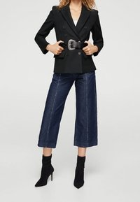 Blazer negro de doble botonadura con solapas de pico, usado sobre una blusa blanca, combinado con pantalones cortos de denim oscuro de pierna ancha y botas de tacón de aguja negras hasta el tobillo.