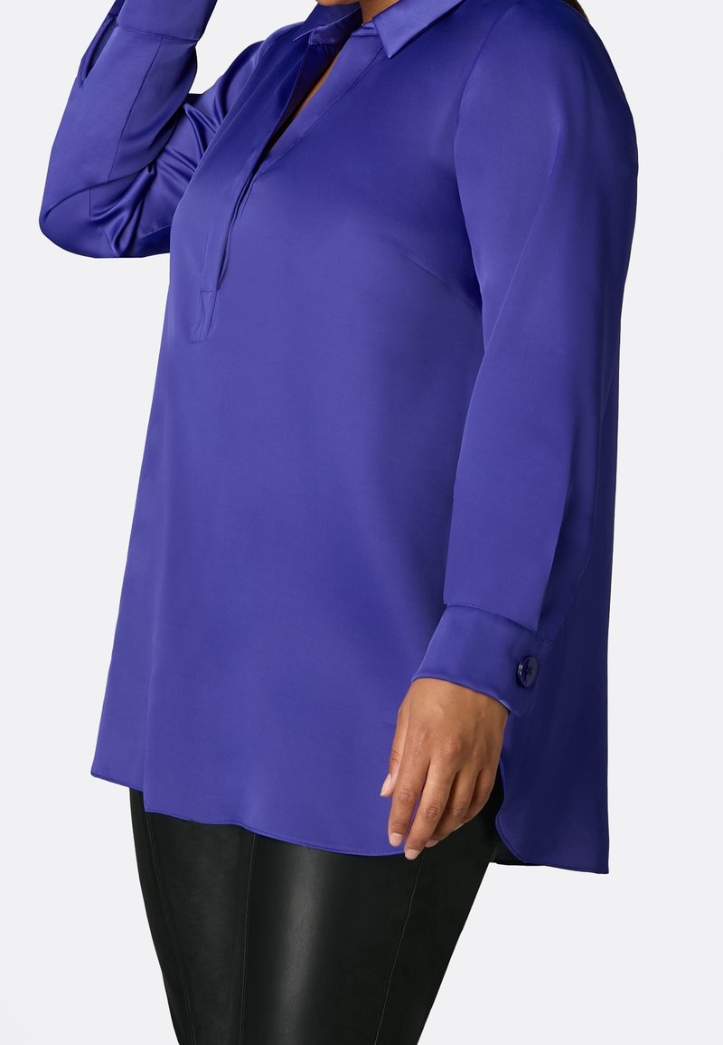 Långärmad lila blus i mjukt tyg, med krage med knäppning och sidoklyftor, tillsammans med svarta byxor i konstläder.