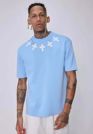 Un homme tatoué porte une chemise bleu clair à manches courtes avec des motifs de croix blanches près du col et un pantalon blanc, debout devant un fond uni.