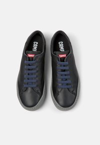 Sneaker in pelle nera con suole in gomma grigia, lacci blu e un'etichetta rossa del marchio. Texture liscia, forma arrotondata, design minimalista.