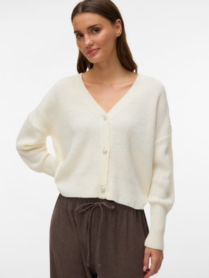 Moteris dėvi kreminės spalvos V formos megztinį su dekoratyvinėmis sagutėmis ir laisvas rudas kelnes su raišteliu, stovinti viena ranka remiasi į klubą.