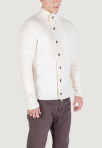 Hombre vistiendo un chaleco de botones color crema con puños y dobladillo acanalados, combinado con pantalones marrón oscuro, de pie contra un fondo claro.