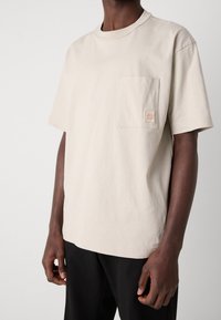 Beige bomull t-shirt med rund halsringning, korta ärmar och en vänster bröstficka med en liten märkeslogotyp. Mjuk textur.