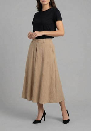 Femme portant un haut noir à manches courtes, une jupe midi beige en ligne A avec poches et des chaussures à talons noires, debout devant un fond gris.