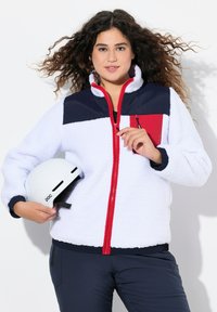 Veste en polaire blanche avec des accents bleu marine et rouge, fermeture éclair à l'avant, col montant et poche poitrine zippée. Modèle tenant un casque blanc.