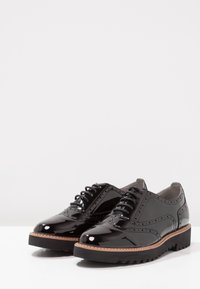 Chaussures oxford en cuir verni noir avec un bout arrondi, des détails en brogue, et une épaisse semelle en caoutchouc noir à finition texturée.