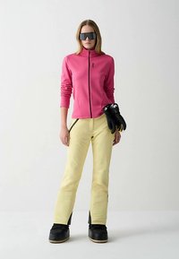 Jakna od flisa ciklama boje s patentnim zatvaračem, žute hlače za snijeg, crne čizme za snijeg i rukavice. Sunčane naočale s ravnim, tamnim staklom. Glatke teksture, ležeran kroj.