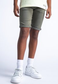 Olivengrønne rullede denimshorts, mellemlangt, parret med hvide sportsko og ankellange hvide sokker, modellens ben synlige.