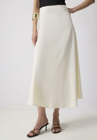 Crème satijnen maxi rok met een vloeiende uitstraling, een zachte textuur en een hoge taille. Gecombineerd met zwarte strappy hakken met een gouden accent.