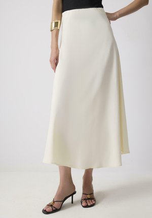 Jupe maxi en satin crème avec un design fluide, une texture lisse et une taille haute. Associée à des talons noirs à lanières avec un accent doré.