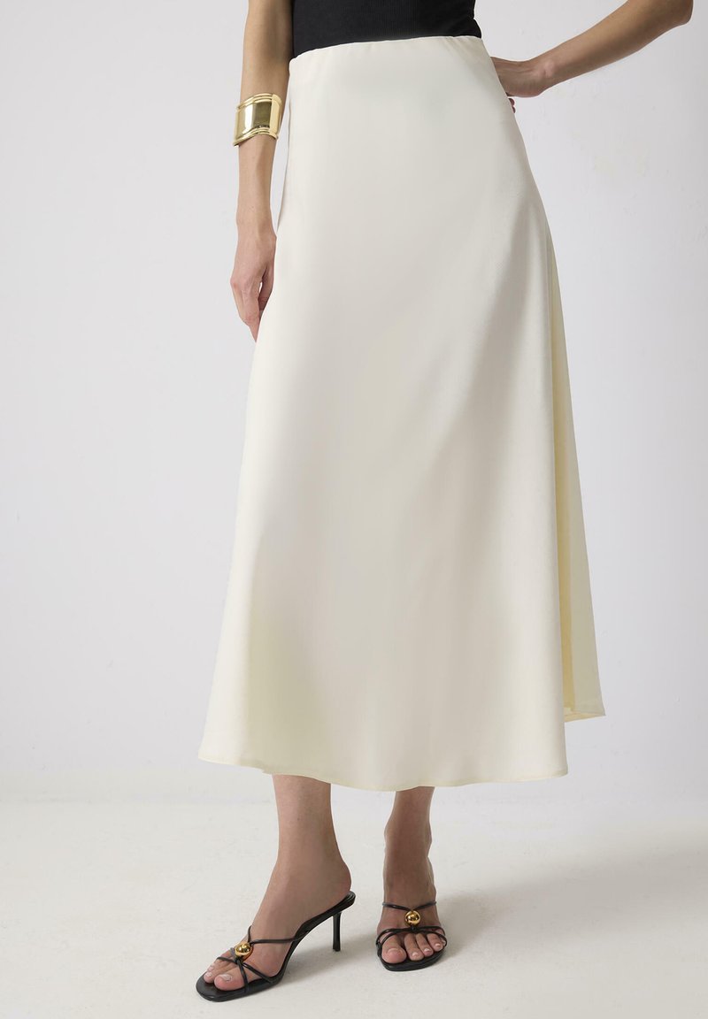 Crème satijnen maxi rok met een vloeiende uitstraling, een zachte textuur en een hoge taille. Gecombineerd met zwarte strappy hakken met een gouden accent.