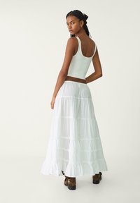 Biely crop top a dlhá sukňa s viacerými vrstvami a volánom na spodnom okraji. Model má na sebe hnedé členkové topánky. Hladká textúra; obidva kúsky sú jednoduchého dizajnu.