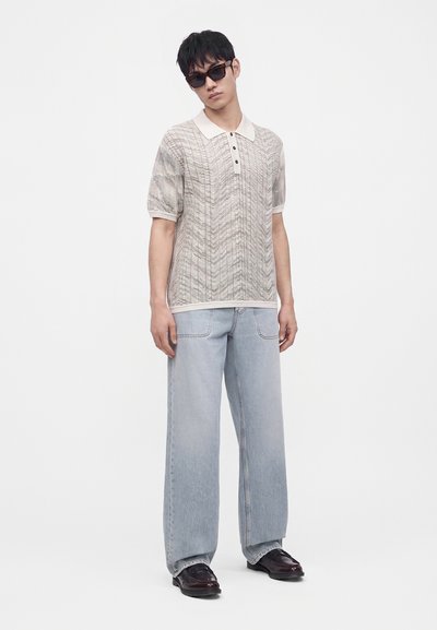 Jeune homme portant des lunettes de soleil foncées, un polo en maille beige à manches courtes, un jean large bleu clair et des chaussures marron foncé, debout sur fond blanc.