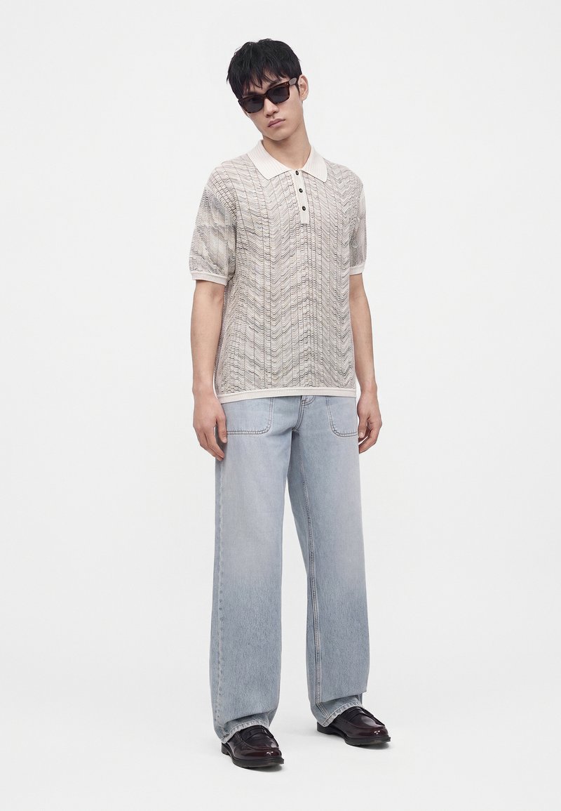 Joven con gafas de sol oscuras, camiseta tipo polo de punto beige de manga corta, vaqueros de pierna ancha azul claro y zapatos marrón oscuro, de pie sobre fondo blanco.