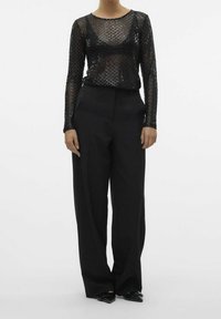 Kvinna klädd i svart genomskinlig långärmad topp med geometriskt mönster, svarta vida byxor och svarta spetsiga skor mot vit bakgrund.