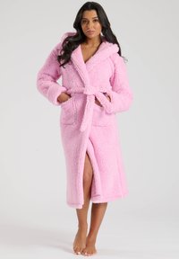 Accappatoio rosa in morbido velluto con cappuccio, maniche lunghe, due tasche frontali e cintura. Presenta una consistenza soffice e pelosa e uno spacco alto fino alla coscia.