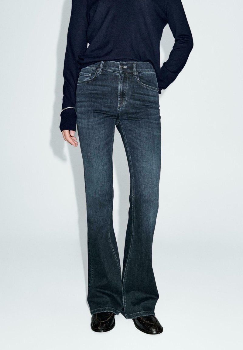 Flared donkerdenim jeans hebben een middelhoog tailleband, een klassieke vijf-pocket stijl en subtiele vervaging voor textuur. Gecombineerd met zwarte schoenen.
