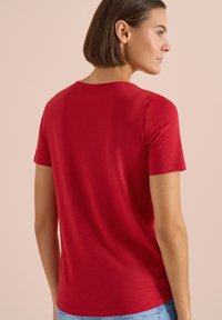 Femme aux cheveux bruns courts vue de dos, portant un t-shirt rouge à manches courtes et un jean bleu clair sur un fond beige.