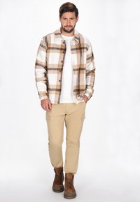 Flanellskjorta i beige och vitt med ett brunt rutmönster, knäppning framtill, bärs över en vit t-shirt, ihop med khaki cargobyxor och bruna kängor.