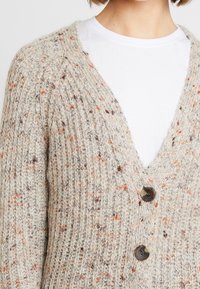Beige pletený kardigan s výstřihem do V, texturovanou látkou, tečkovaným designem a dvěma tmavými knoflíky vpředu. Nesený přes bílé tričko.