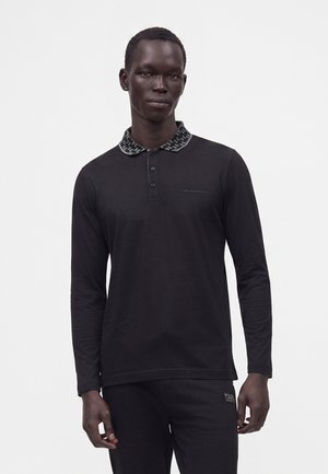 Mann trägt ein schwarzes langärmliges Poloshirt mit gemustertem Kragen und passenden schwarzen Hosen vor hellem einfarbigem Hintergrund.
