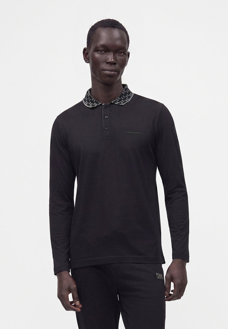Homme portant un polo noir à manches longues avec un col à motifs et un pantalon noir assorti sur un fond clair uni.