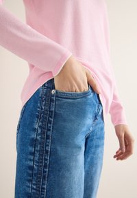 Personne portant un haut à manches longues rose et un jean bleu, la main gauche placée dans la poche avant, sur un fond neutre.