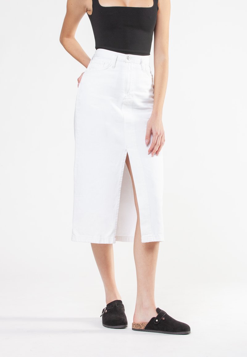 Falda midi de denim blanco con una abertura frontal, cintura alta y lazos para cinturón. Combinada con unos zapatos negros de tipo slip-on con un acento de corcho.