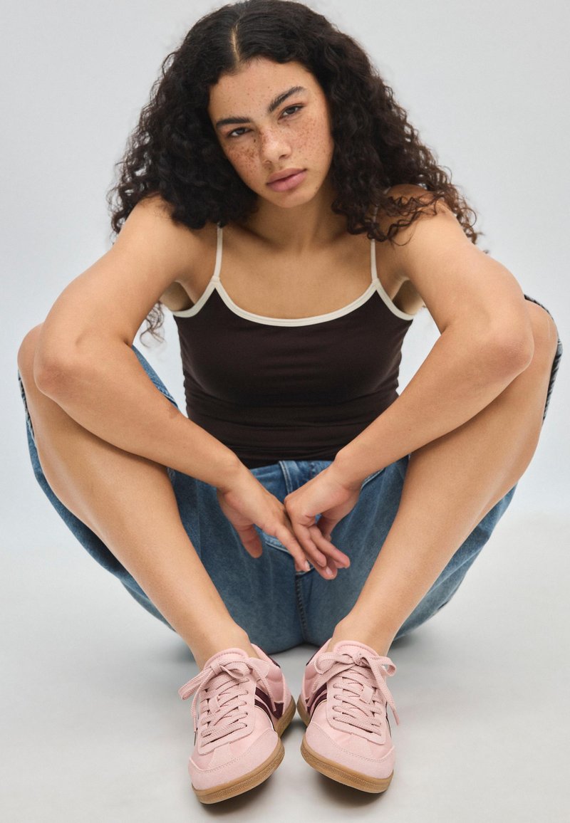 Jeune femme aux cheveux bouclés, taches de rousseur et vêtements décontractés, assise en tailleur, portant des baskets roses et un short en jean.