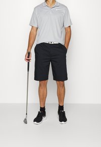 Šedé polo tričko Nike a černé šortky s texturovaným povrchem. Černé boty a ponožky Nike. Drží golfovou hůl. Jednoduchý design.