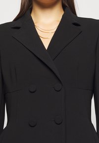 Černý dvouřadý blazer s klopami, který má kulaté knoflíky a strukturované ramena. Hladká, texturovaná tkanina s přizpůsobeným střihem.