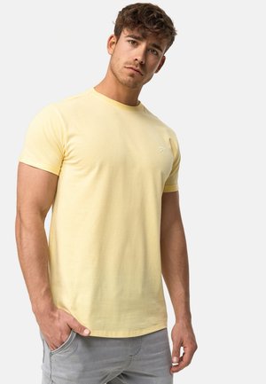 Gele katoenen T-shirt met een ronde hals en korte mouwen, voorzien van een klein wit logo op de linkerborst, gedragen met grijze broek.