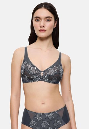 Moteris, dėvinti tamsiai pilką permatomą liemenėlę ir derančius aukštai iškilusius apatinius su baltais gėlių motyvais, stovi prieš paprastą foną.