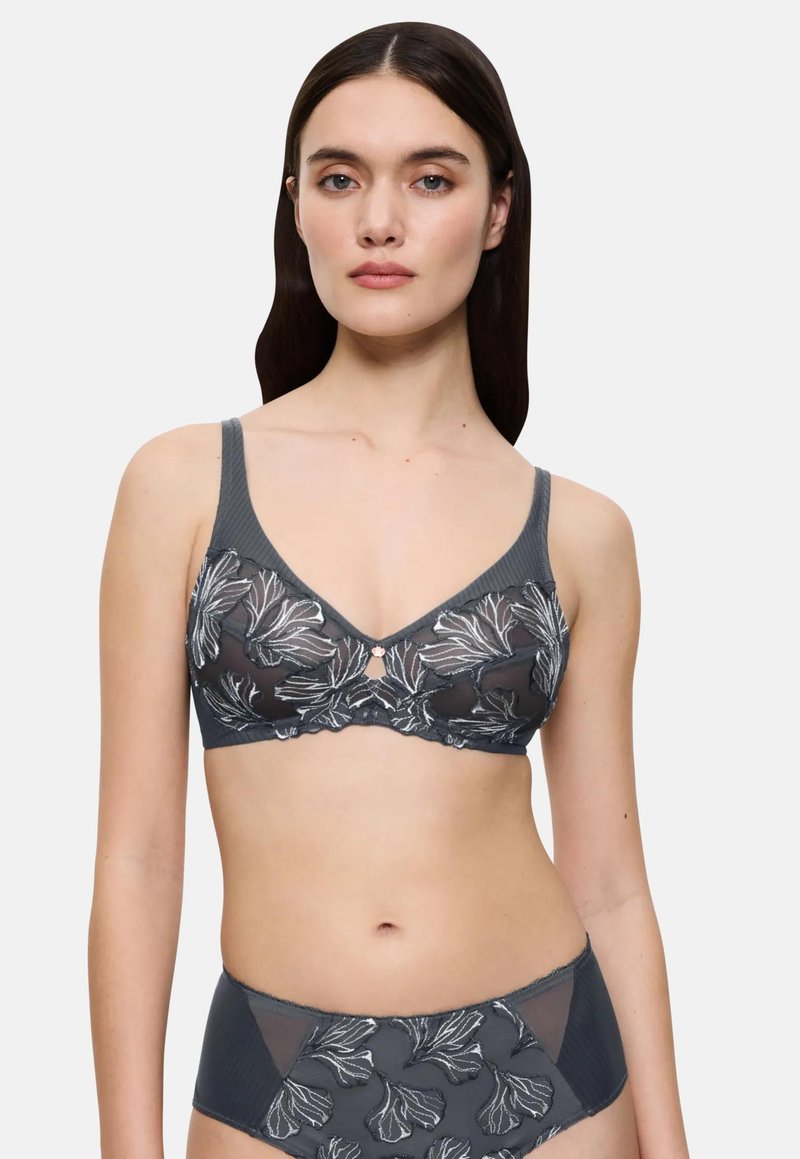 Sieviete, tērpusies tumši pelēkā caurspīdīgā krūšturī un atbilstošās augsta vidukļa biksītēs ar baltu ziedu izšuvumu, stāv pretī vienkāršai fona.