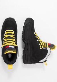 Černé nubukové turistické boty s robustní černou podrážkou, žlutými tkaničkami a vzorovaným jazykem. Obsahují bílý akcent poblíž paty.