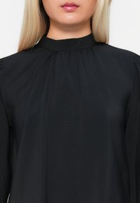Blusa nera a maniche lunghe con collo alto, tessuto morbido, dettagli arricciati sul collo e una sottile texture. Nessun motivo o accentuazione visibile.