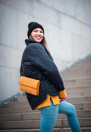 Černá péřová bunda, hořčicový pletený svetr, světle modré skinny džíny, černá pletená čepice a oranžová taška přes rameno. Model pózuje na schodech.