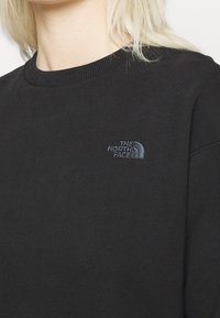Gros plan sur une personne portant un sweat-shirt noir avec un petit logo brodé "THE NORTH FACE" sur le côté gauche de la poitrine.
