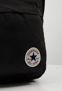 Sac à dos en toile noir avec un patch rond comportant une étoile bleue et le texte "Converse Chuck Taylor All Star" en rouge et blanc.
