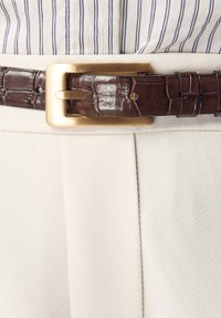 Brunt bälte i krokodilmönstrat läder med spänne av stort rektangulärt guld, bärs med krämfärgade byxor och en randig skjorta.