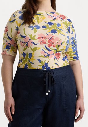 Frau trägt ein cremefarbenes Oberteil mit rosa, blauen und gelben Blumenmuster, kombiniert mit hoch taillierter marineblauer Hose, die in der Taille gebunden ist.