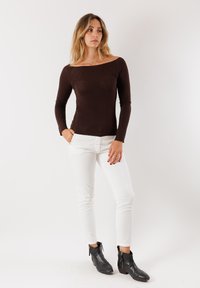 Top lungo marrone con design off-the-shoulder, abbinato a pantaloni bianchi slim-fit e stivaletti neri alla caviglia con zip.