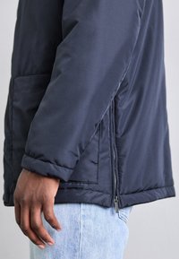 Sido vy av en person som bär en marinblå vadderad jacka med dragkedjedetalj över ljusblå jeans, handen avslappnad vid sin sida.