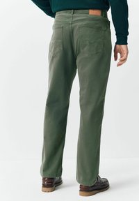 Mees, kes kannab rohelisi sirge säärega pükse, pruunist nahast saapaid ja tumerohelist pikkade varrukatega särki, seisab tavalise valge tausta vastu.