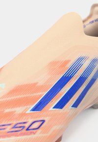 Primer plano de una bota de fútbol color durazno con franjas azules de Adidas y texto "F50" en azul en el lateral.