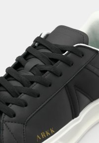 Sorte lærersneakers med flate svarte lisser, hvit såle og minimalistisk design. Har gullbranding på siden og teksturerte detaljer.