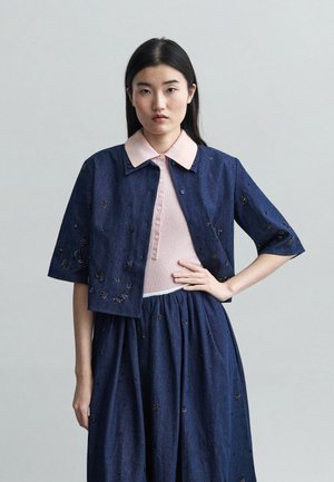 Jeune femme aux longs cheveux noirs portant une veste et une jupe brodées bleu marine par-dessus une chemise rose à col, posant devant un fond gris uni.